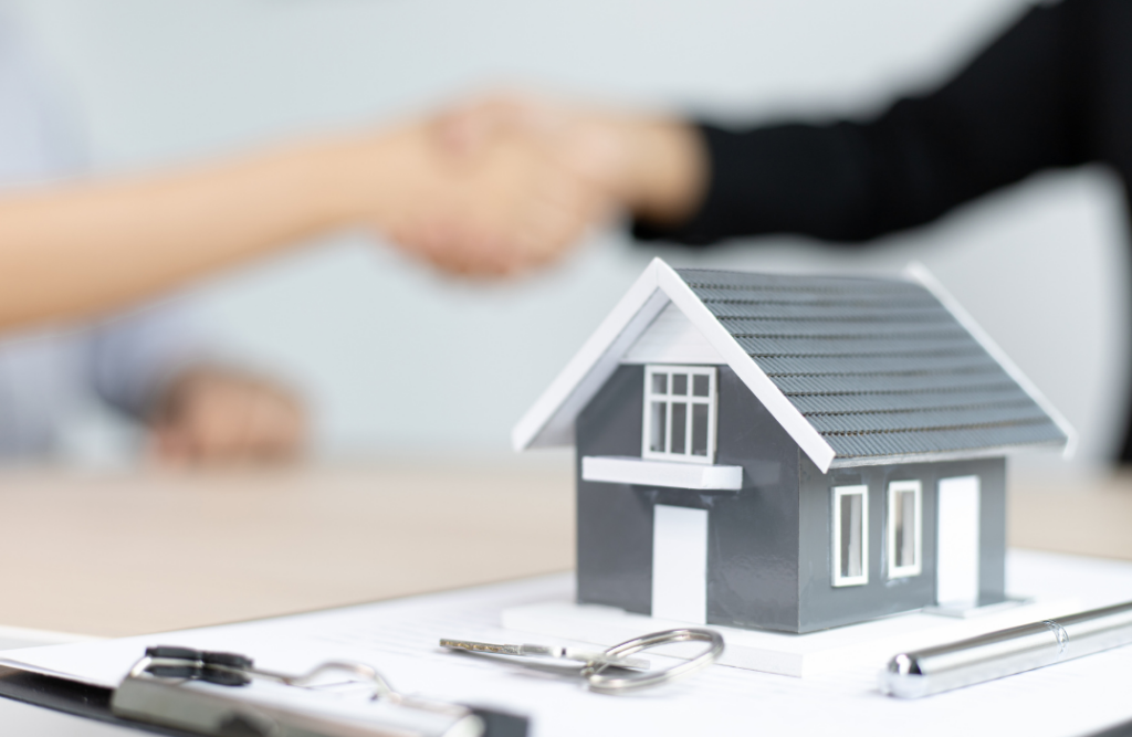 Miniature house model on a desk alongside keys and a pen, with two individuals shaking hands in the background, symbolizing a property agreement.