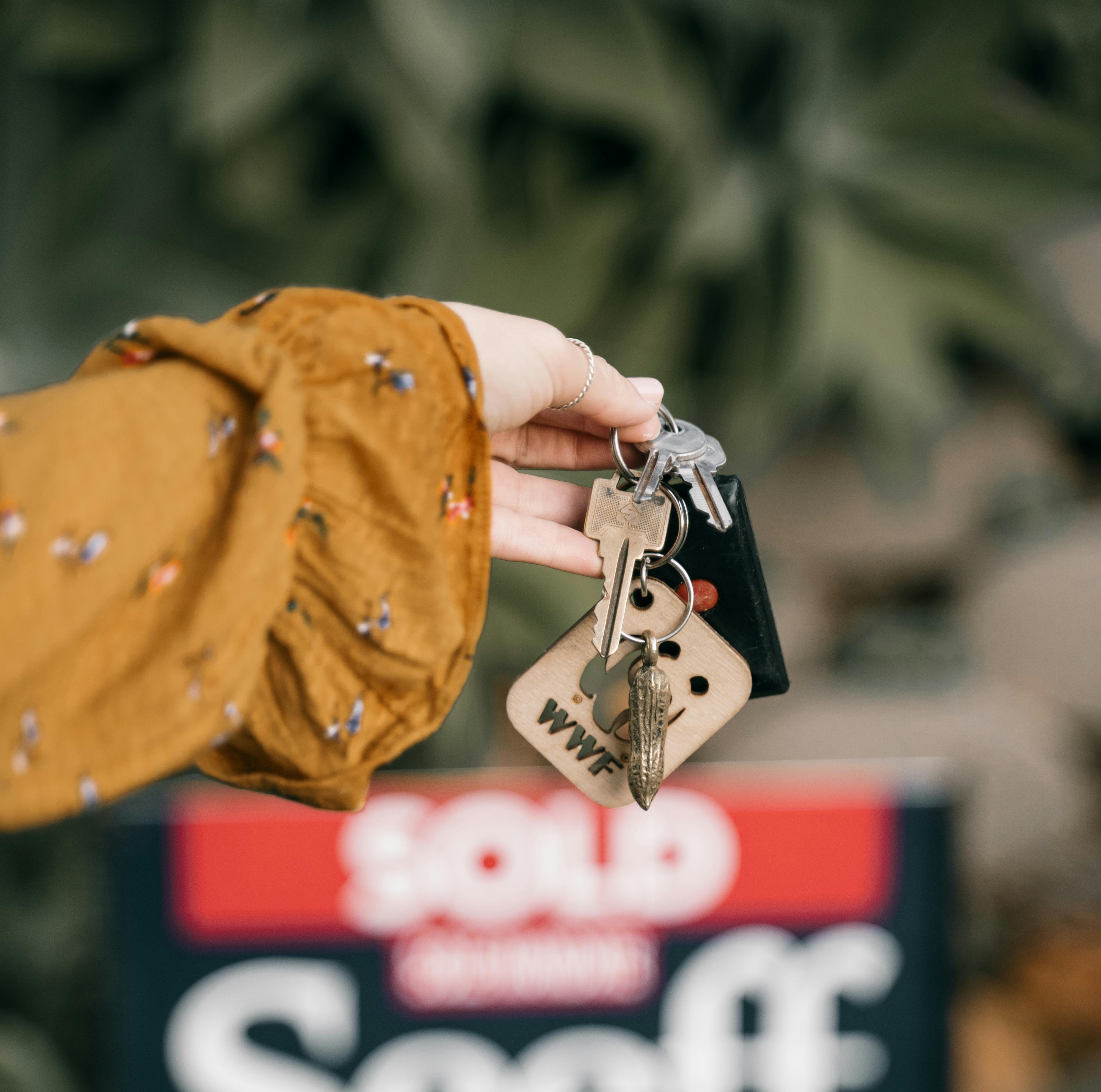 A person holding house keys in front of a sold property sign, illustrating tips on how to manage and list houses online.